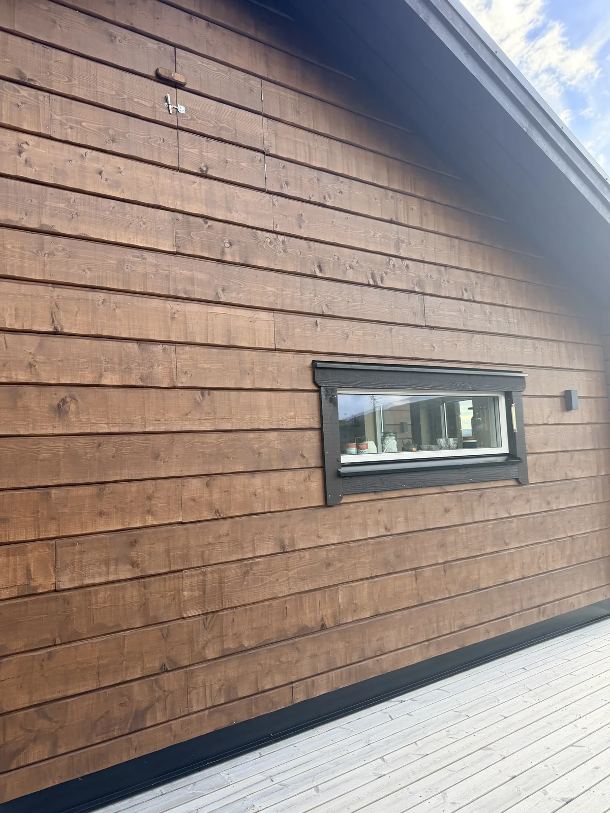 Brunlaserad fasad i liggande panel med ett litet fönster med svart karm och trädäck nedanför