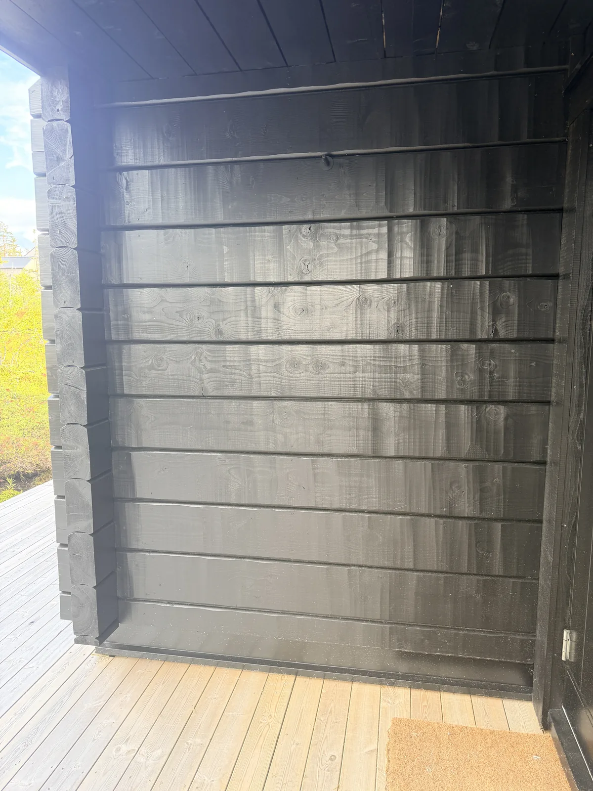 Svartlaserad panelvägg under utskjutande tak vid entré, med dörrmatta på ljust trädäck